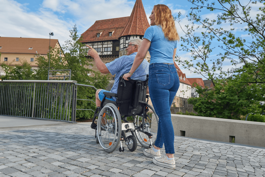 Das Bild zeigt eine junge Frau von hinten, die einen älteren Mann im Rollstuhl auf einem Weg mit Pflastersteinen schiebt. Im Hintergrund ist eine Burg zu sehen. Am Rollstuhl befindet sich hinten eine Schiebehilfe V-MAX mini, welche über das Antriebsrad beim Schieben unterstützt.