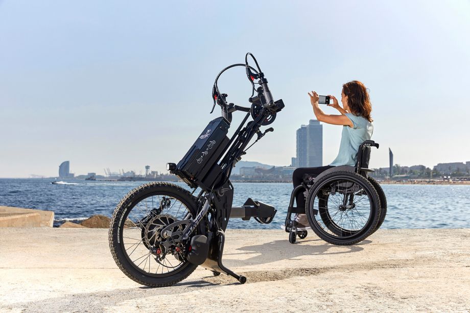 Im Hintergrund des Bildes ist die junge Frau zu sehen, wie sie mit dem Smartphone das Meer fotografiert. Im Vordergrund steht das Rollstuhl-Handbike Hybrid 2, welches sie von ihrem Rollstuhl abgekoppelt hat. 