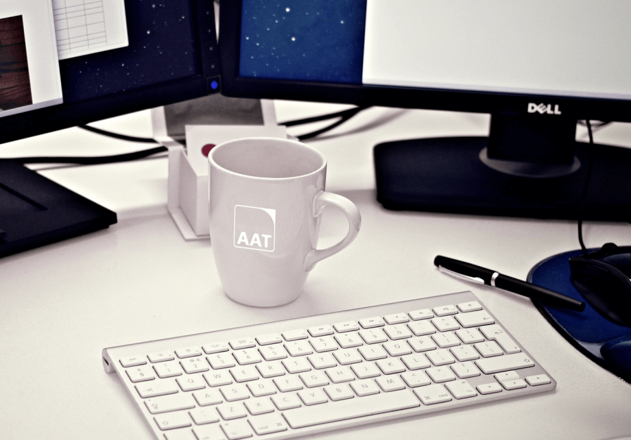 Auf dem Bild ist ein Arbeitsplatz zu sehen, auf dem eine weiße Porzellantasse mit eingraviertem AAT-Logo zu sehen ist. Im Hintergrund sind zwei Bildschirme zu sehen und vor der Tasse liegt eine Tastatur. 