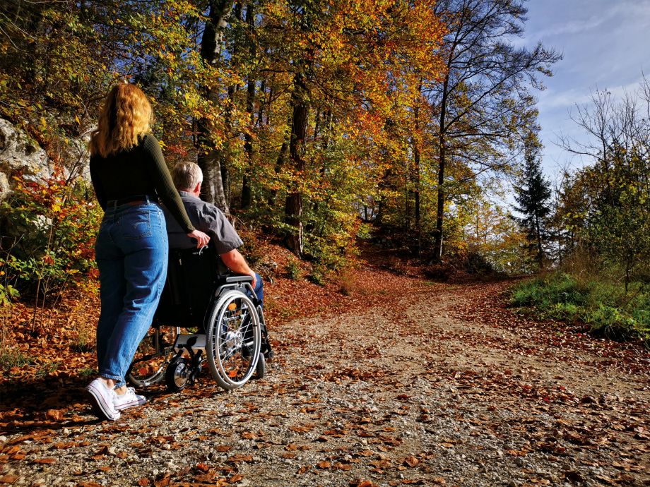 Das Bild zeigt eine junge Frau von hinten, die einen älteren Mann im Rollstuhl einen Schotterweg im Wald hinaufschiebt. Die Bäume sind bunt und es liegt Laub auf dem Weg. Am Rollstuhl befindet sich hinten eine Schiebehilfe V-MAX mini, welche über das Antriebsrad beim Schieben unterstützt.