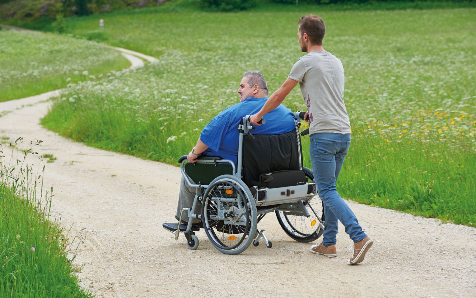 Das Bild zeigt einen schlanken Mann mittleren Alters, der einen anderen Mann in einem XXL-Rollstuhl auf einem Feldweg schiebt. Der Rollstuhlinsasse hat ein sehr hohes Körpergewicht, weshalb sich hinten am Rollstuhl eine Brems- und Schiebehilfe V-MAX+ befindet, die die Begleitperson beim Schieben und Bremsen des Rollstuhls unterstützt. 