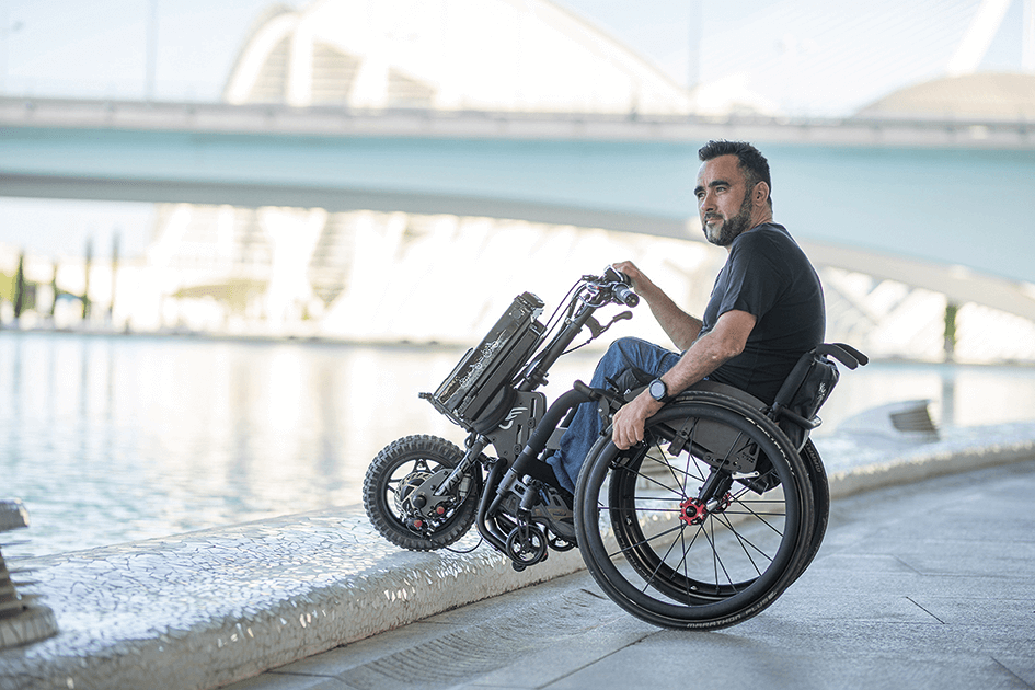 Das Bild zeigt einen Mann im Rollstuhl, der ein kompaktes Rollstuhl-Zuggerät verwendet. Er steht vor einem modernen Gebäude mit angelegtem Teich nur auf den hinteren Rollstuhlrädern.