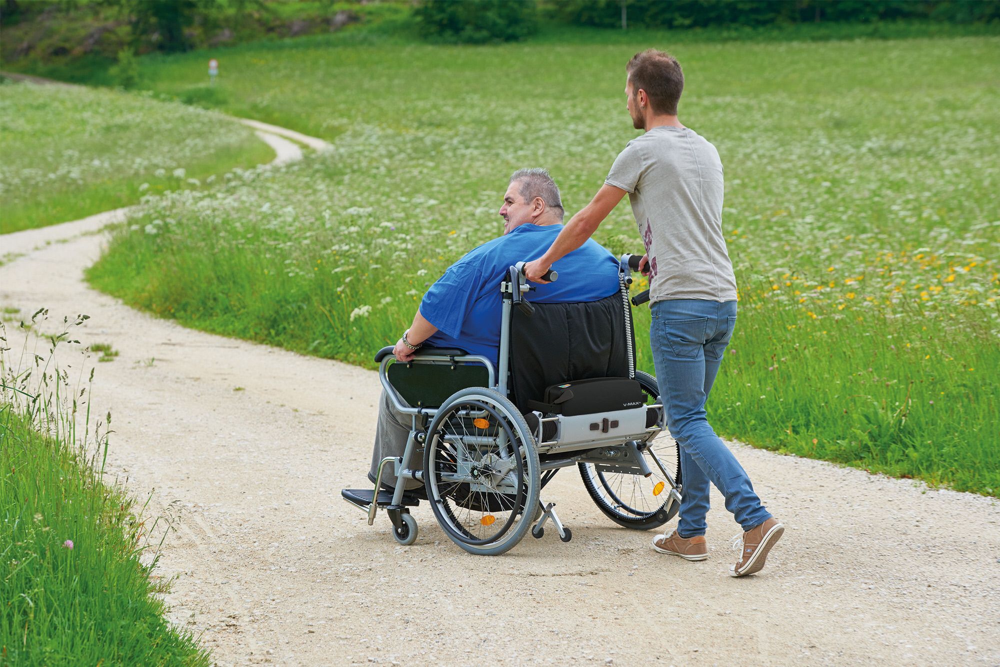 Das Bild zeigt einen adipösen Mann, der in seinem XXL-Rollstuhl von einem anderen Mann geschoben wird. Um die Begleitperson zu entlasten, wird eine Schiebe- und Bremshilfe für den Rollstuhl genutzt.