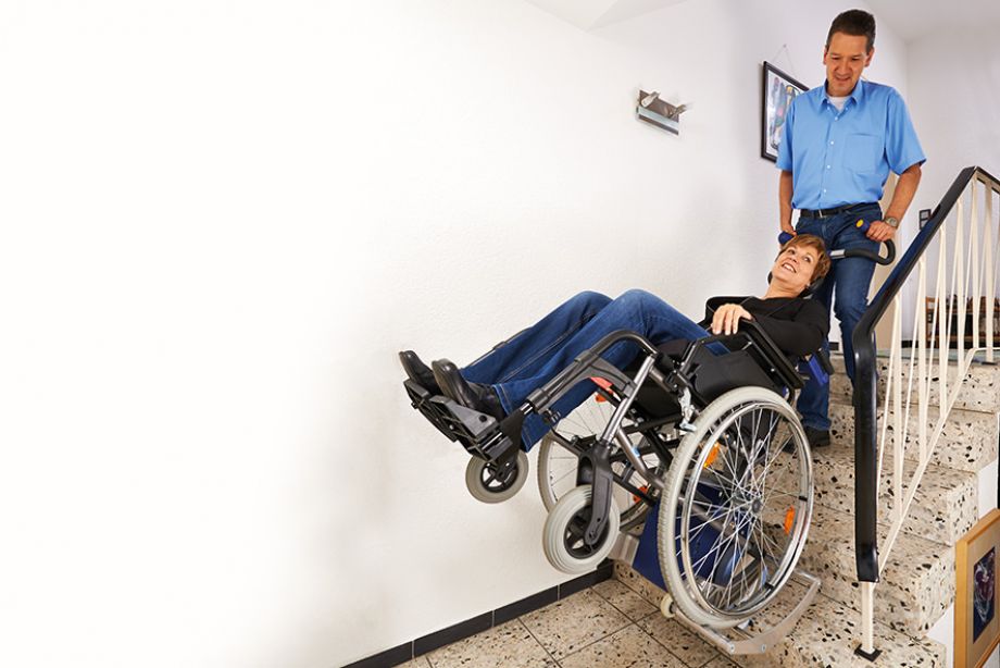 Das Bild zeigt einen Mann mittleren Alters, der eine Frau im Rollstuhl über Treppen innerhalb eines Hauses befördert. Der Rollstuhl wurde hierzu auf die Aufnahmeschuhe des Treppensteiger s-max in Kombination mit der Universalaufnahme SDM7 geschoben. 