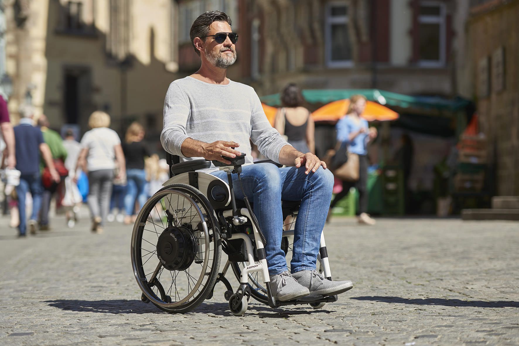 Das Bild zeigt einen Mann mit Sonnenbrille, der an einem sonnigen Tag mit seinem Rollstuhl durch die Stadt fährt. Er nutzt einen Rollstuhl-Zusatzantrieb mit Joystick.