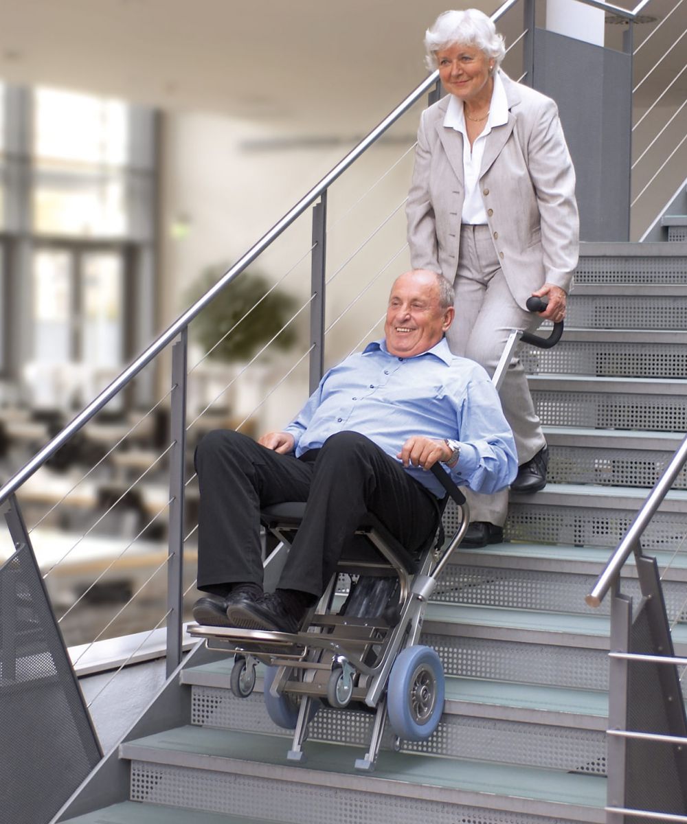 Das Bild zeigt einen Mann mittleren Alters, der von einer Frau mithilfe eines Treppensteigers die Treppen hinaufbefördert wird. 