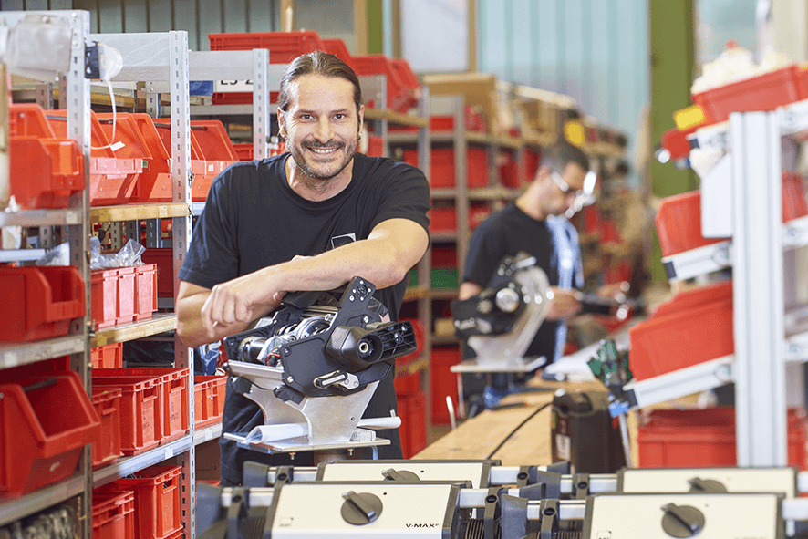 Das Bild zeigt einen Mitarbeiter, der in der Produktionshalle von AAT an seinem Arbeitsplatz steht. Er hat einen Arm auf der Schiebehilfe, die er selbst gebaut hat, abgelegt.