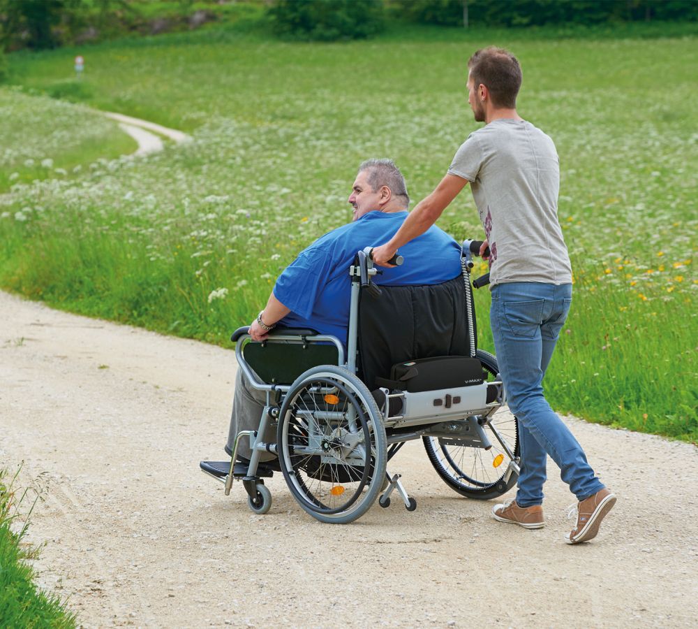 Das Bild zeigt einen schlanken Mann, beim Schieben eines Mannes im Rollstuhl. Dieser hat starkes Übergewicht und zur Unterstützung wird eine Schiebe- und Bremshilfe V-MAX+ verwendet.