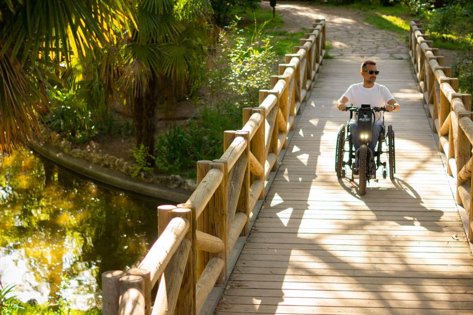 Auf dem Bild ist erneut der Mann mit Sonnenbrille zu sehen, der an einem sonnigen Tag mit seinem Rollstuhlzuggerät BATEC SCRAMBLER 2 durch einen angelegten Park fährt. Auf diesem Bild fährt er über eine breite Holzbrücke.