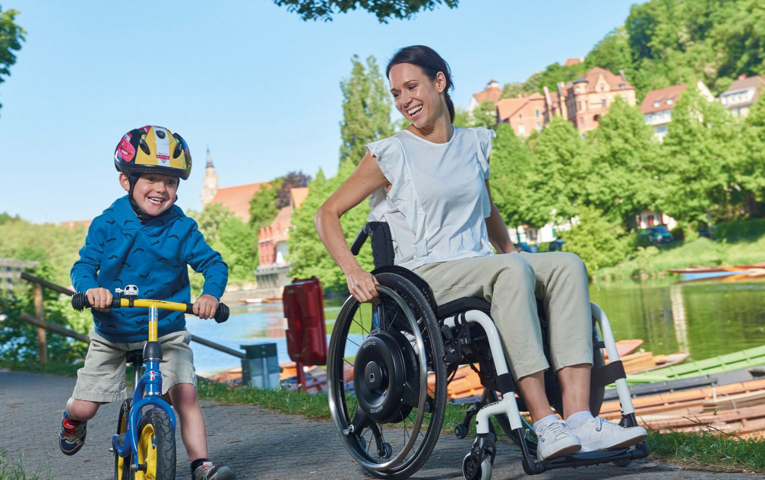 The picture shows a woman sitting in a wheelchair and riding along a river with her son. She is propelling the wheelchair herself with her arms. An AAT SERVO residual force booster is attached to the wheelchair to provide power assistance for the woman. 
