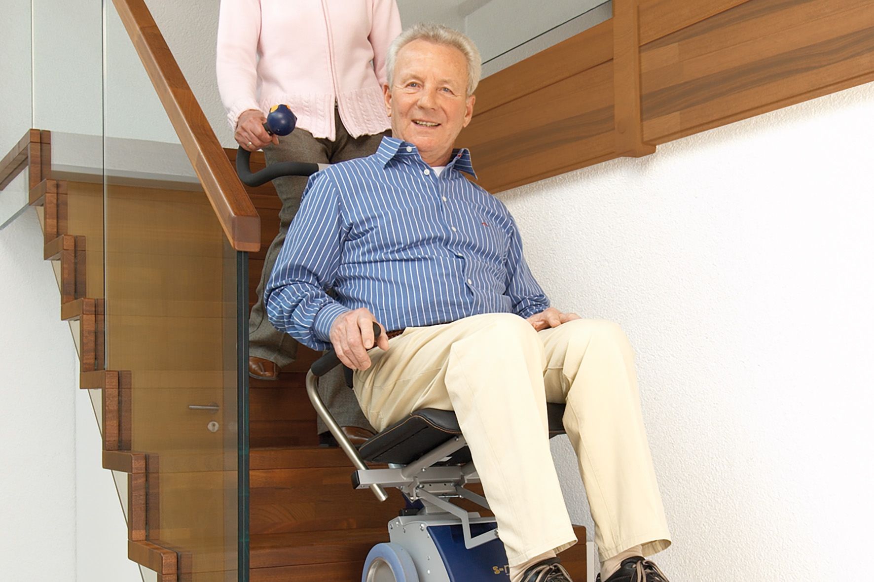Das Bild zeigt einen älteren Herren, der in einem Treppensteiger mit integrierter Sitzeinheit von einer Frau über eine Treppe transportiert wird.