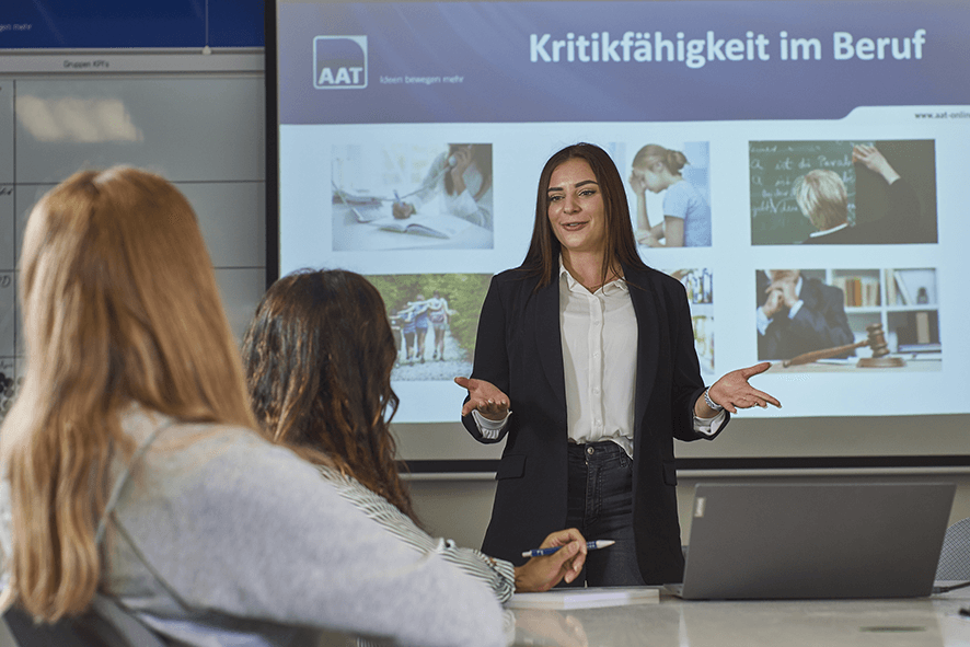 Das Bild zeigt eine junge Auszubildende, die vor einer Gruppe Kollegen einen Vortrag über Kritikfähigkeit im Beruf hält. Im Hintergrund ist ihre Präsentation zu sehen.