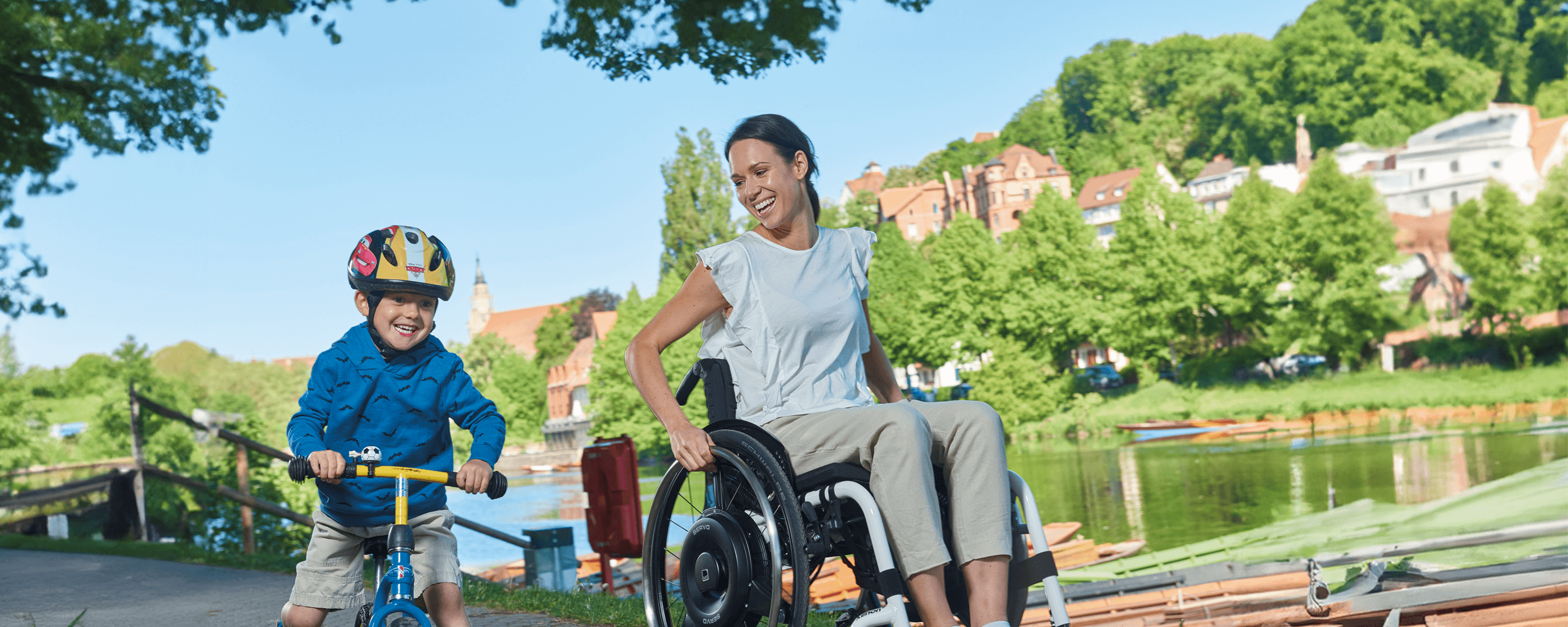 Eine junge Frau im Rollstuhl nutzt einen schwarzen Radnabenantrieb zur Restkraftverstärkung und fährt mit ihrem Sohn auf einem Cityroller an einem Fluss entlang.