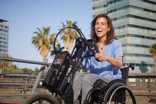 Das Bild zeigt eine junge Frau im Rollstuhl, die gemeinsam mit ihrem Sohn auf einem Laufrad einen Ausflug an einem Fluss macht. Beide haben viel Spaß. Die Frau nutzt einen Restkraftverstärkenden Antrieb für Ihren Rollstuhl.