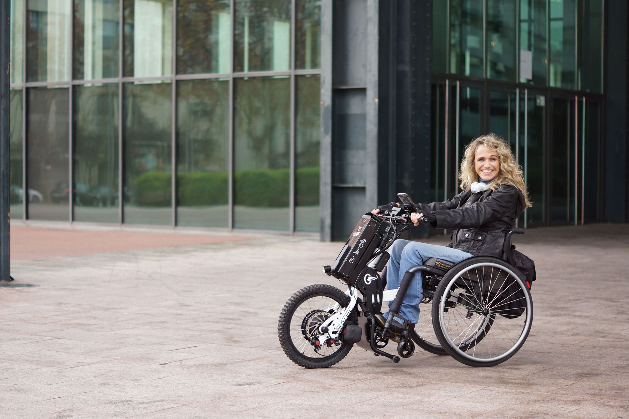Das Bild zeigt eine blonde Frau im Rollstuhl, die ein Rollstuhl-Zuggerät verwendet. Sie fährt an einem modernen Bürogebäude vorbei und lächelt in die Kamera. 