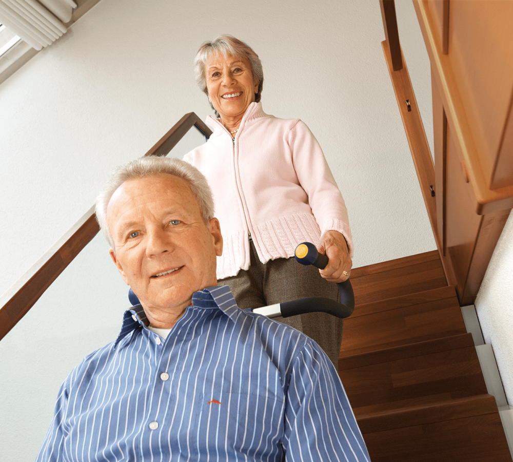 Das Bild zeigt einen Mann mittleren Alters, der von einer Frau in seinem Rollstuhl mithilfe eines Treppensteigers die Treppen hinaufbefördert wird. Beide lächeln in die Kamera.
