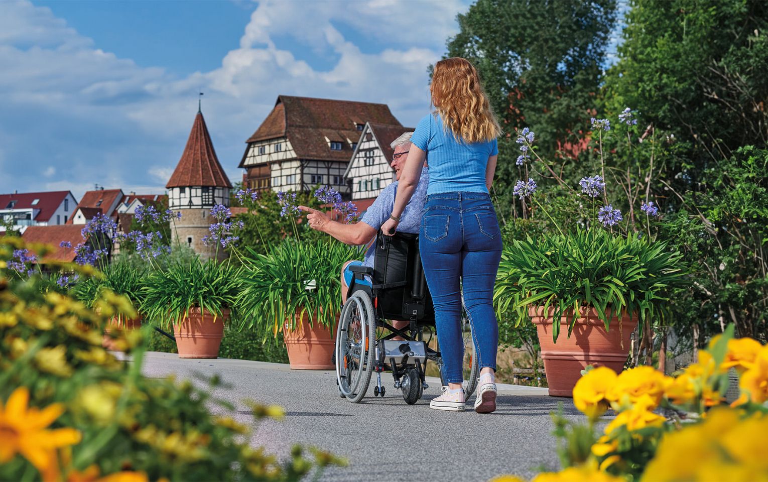 Das Bild zeigt eine jüngere Frau, die einen älteren Mann im Rollstuhl durch eine blühende Stadt schiebt. Im Hintergrund ist eine Burg zu sehen. Am Rollstuhl befindet sich eine AAT Schiebehilfe, die die Frau beim Schieben des Rollstuhls entlastet. 