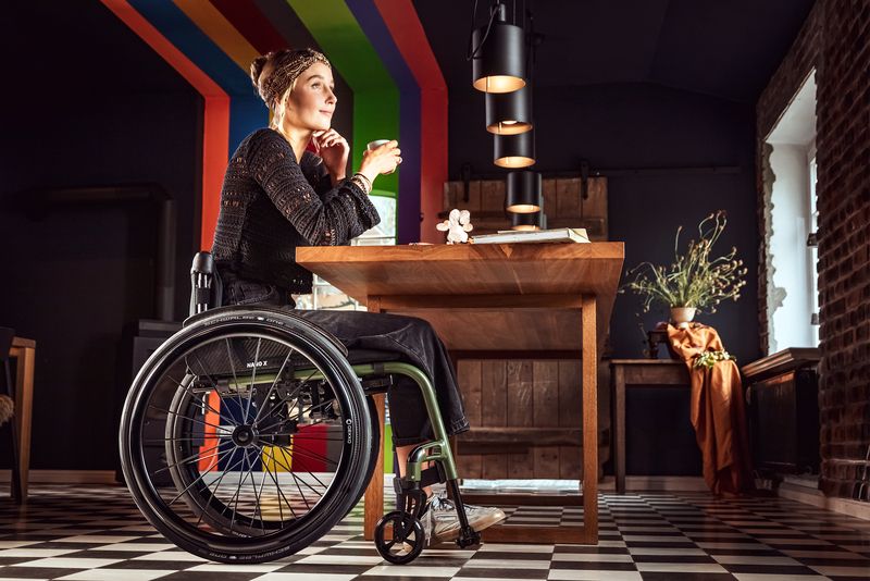 Das Bild zeigt eine junge Frau, die in einem modernen Café sitzt und ein Heißgetränk trinkt. Mit ihrem modernen, grün-schwarz gehaltenen Aktivrollstuhl kann sie problemlos unter den Tisch fahren. Die Tischkante stellt kein Hinderniss dar. 
