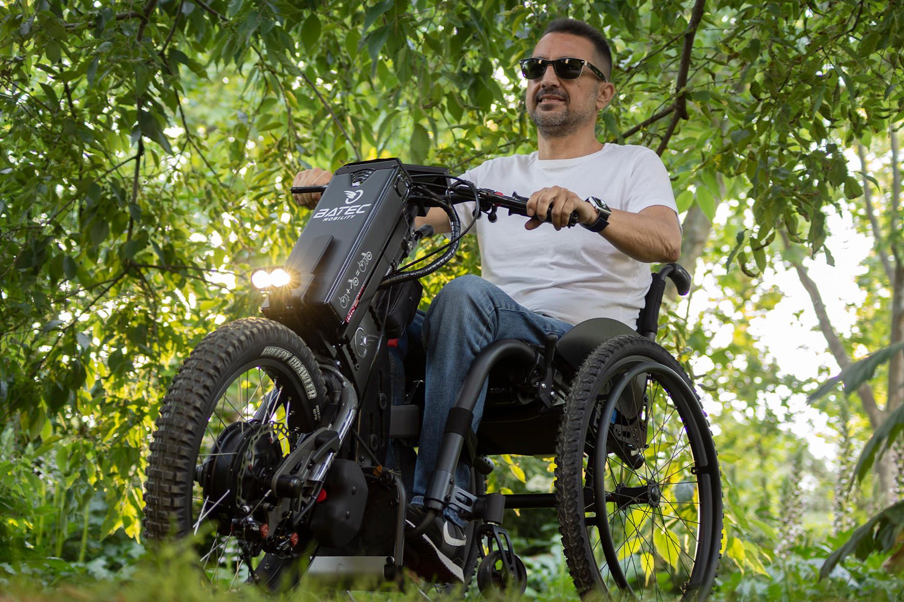 Das Bild zeigt einen Mann im Rollstuhl, der mit Sonnenbrille im Wald unterwegs ist. Er nutzt hierzu ein Rollstuhl-Zuggerät im Offroad-Look.