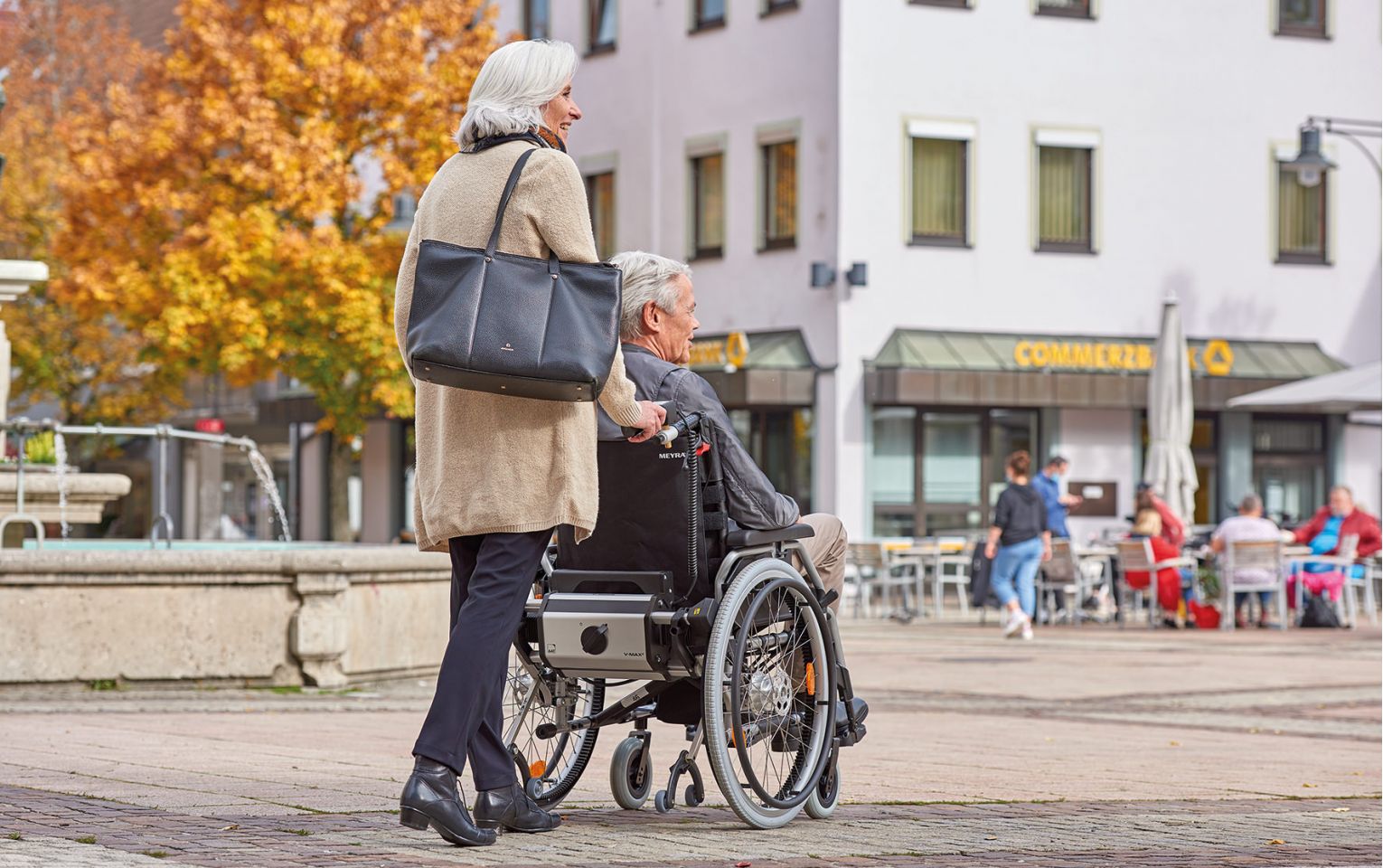 The picture shows an elderly woman pushing an elderly man in his wheelchair through a city center with cobblestones. The wheelchair is fitted with a V-MAX² pushing and braking aid from AAT Alber Antriebstechnik, which supports the woman when pushing and braking the wheelchair and thus offers greater safety.